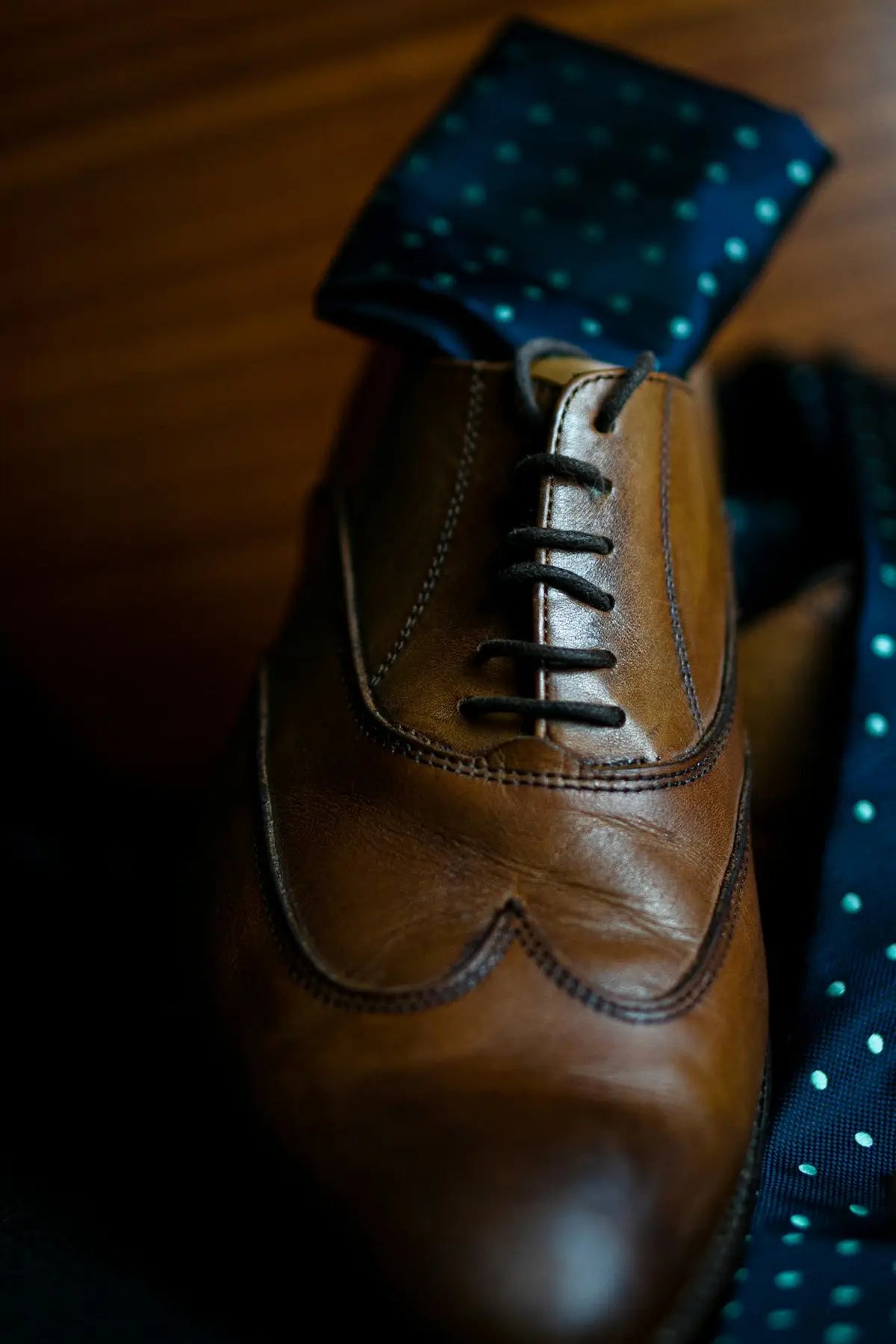 Brown leather wingtip oxford shoe with black laces and a dark blue polka dot pocket square.